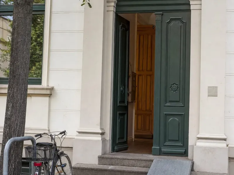 Geöffnete grüne Eingangstür eines hellen Altbaus mit Rundbogen, davor eine graue Rampe über die Stufen, links ein Baum und ein abgestelltes Fahrrad.