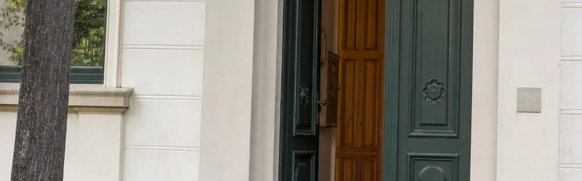 Geöffnete grüne Eingangstür eines hellen Altbaus mit Rundbogen, davor eine graue Rampe über die Stufen, links ein Baum und ein abgestelltes Fahrrad.