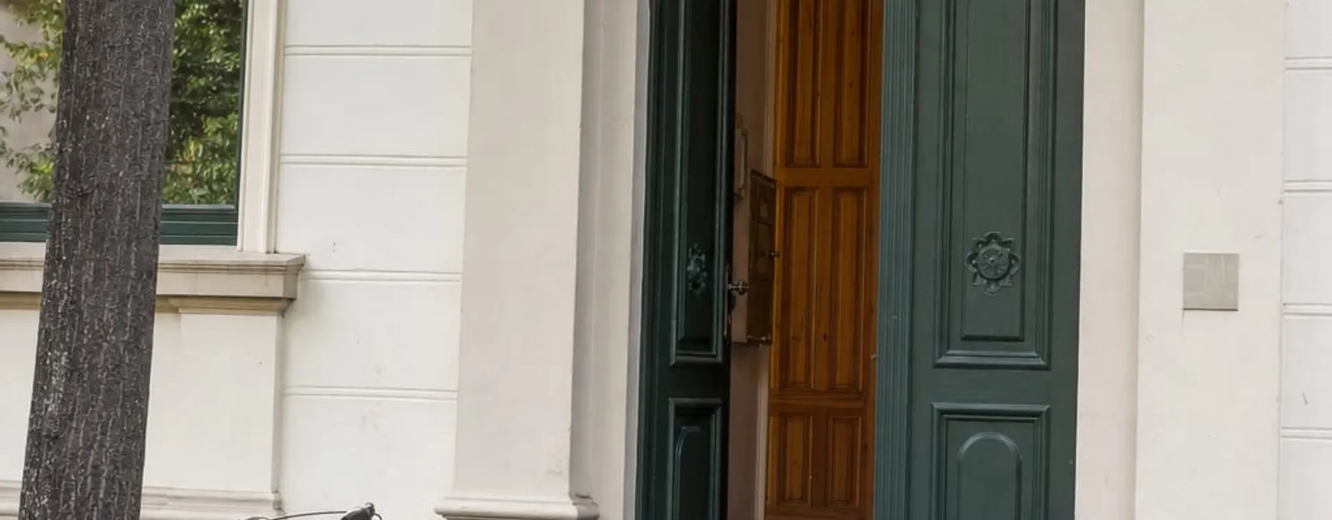 Geöffnete grüne Eingangstür eines hellen Altbaus mit Rundbogen, davor eine graue Rampe über die Stufen, links ein Baum und ein abgestelltes Fahrrad.