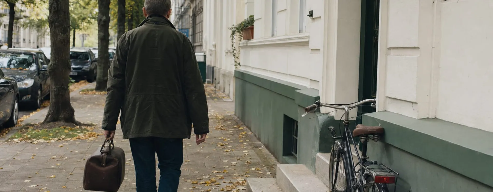 Älterer Mann in olivgrüner Jacke geht mit brauner Ledertasche eine herbstliche Altbau-Straße entlang, rechts ein angelehntes Fahrrad.