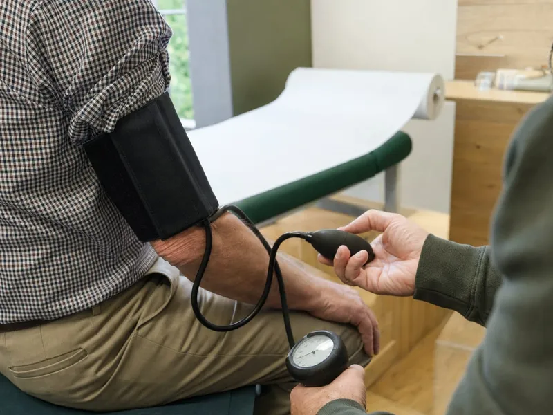 Blutdruckmessung im Behandlungsraum: Eine Person hält den Messballon, am Oberarm des sitzenden Patienten liegt eine schwarze Manschette.